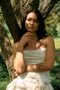 Kıvırcık saçlı genç bir kadın ılık yaz güneşinin altında dalgalı beyaz bir elbise içinde poz veriyor..