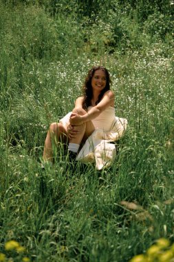 A joyful young woman with curly hair poses in a white dress amidst green grass.