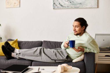 Genç yakışıklı bir adam rahat bir şekilde kanepede uzanıyor, rahat yaşam alanında içkisini yudumluyor..