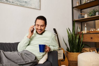 Konforlu bir oturma odasında dinlenen genç bir adam, içkisini yudumlarken telefonda mutlu bir şekilde sohbet ediyor..