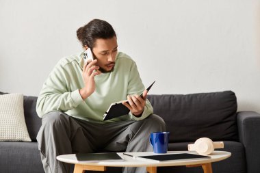 Evde telefonla konuşan genç bir adam, rahat bir kanepede dinlenirken defterine notlar alıyor..