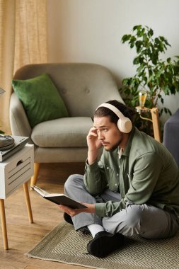 Genç yakışıklı bir adam yerde oturuyor, rahat yaşam alanında kulaklık takıp kitap okuyor..