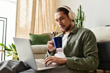 Genç bir adam evde oturuyor, kahvesini yudumluyor ve dikkatle laptopuyla çalışıyor..