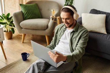 Yakışıklı genç adam yerde oturuyor, dizüstü bilgisayarıyla meşgul, ev konforuyla çevrili..