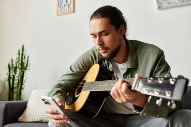 Yakışıklı genç adam evdeki kanepede oturup gitarını tıngırdatıyor ve telefonunu kontrol ediyor..