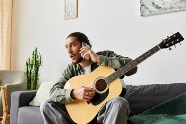 Genç bir adam rahat bir yaşam alanında gitarını çalarken telefonda hevesle konuşuyor..