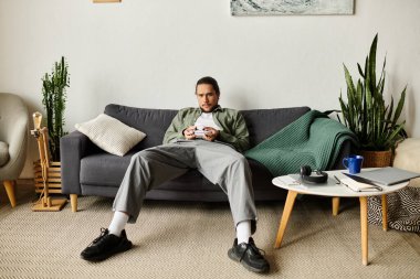 Genç bir adam, şık bir evde video oyunlarına dalmış bir şekilde kanepede dinleniyor..