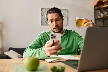 Genç yakışıklı bir adam dizüstü bilgisayarla masada oturuyor, çayını yudumluyor ve telefonunu kontrol ediyor..