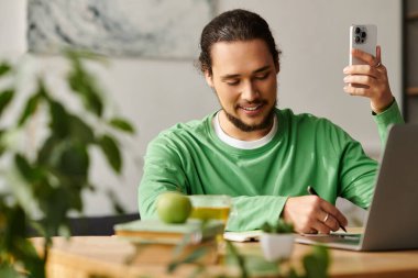 Yakışıklı genç adam deftere yazarken gülümsüyor ve evde akıllı telefon kullanıyor..