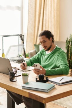Genç bir adam evindeki modern bir masada oturuyor, notlar yazarken akıllı telefonuna odaklanıyor..