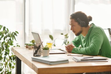 Yeşil kazaklı odaklanmış genç adam rahat bir iş yerinde dizüstü bilgisayarıyla meşgul oluyor..