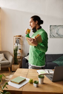 Yeşil gömlekli genç bir adam evde durmuş çayını yudumluyor ve rahat bir ortamda kitap tutuyor..