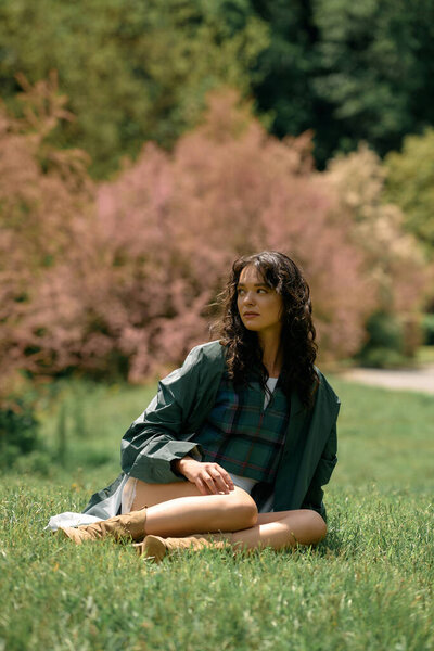 A young woman sits elegantly on the grass, modeling her stylish raincoat in a vibrant setting.