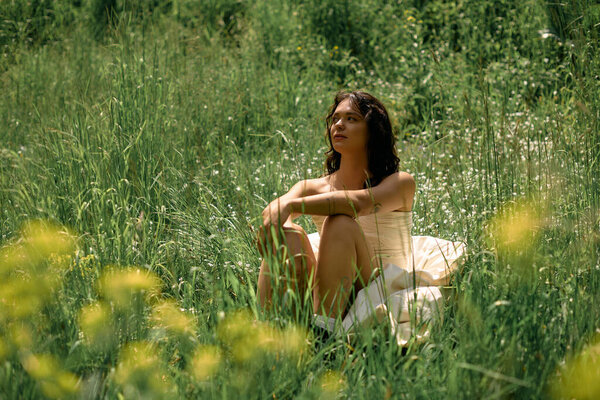 A young woman in a flowing white dress sits peacefully surrounded by lush greenery and wildflowers.