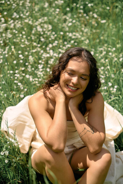 A young woman with curly hair sits gracefully in a field of wildflowers, smiling joyfully.