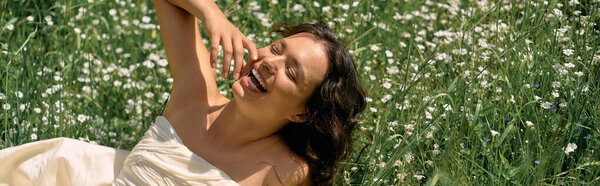 A young woman with curly hair smiles brightly while lounging in a meadow filled with flowers, banner