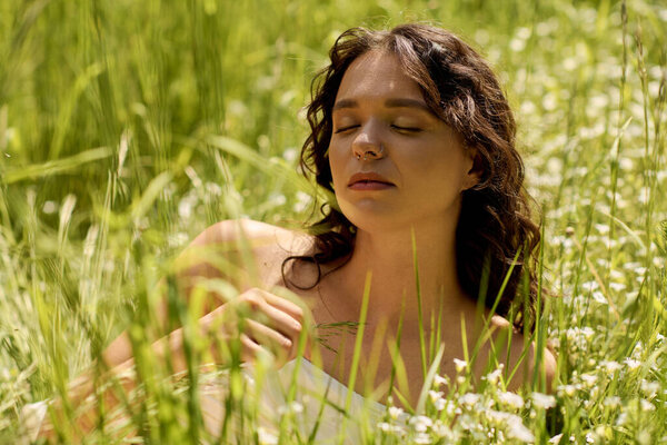 Curly-haired woman relaxes in sunlight, surrounded by green grass and flowers on a warm day.