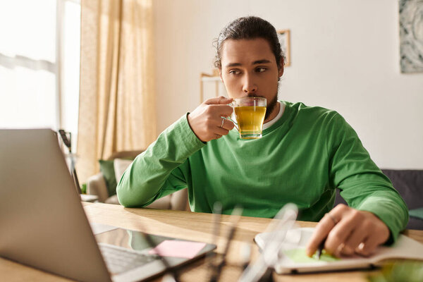 Молодой красивый мужчина сидит дома и задумчиво потягивает чай за своим столом..