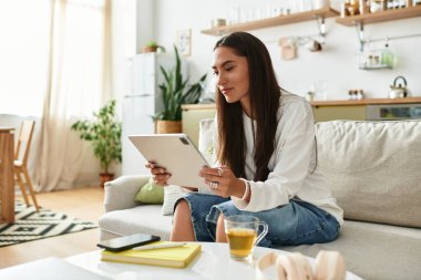 Genç kadın evinde dinleniyor, sıcak bir atmosferde çayını yudumlarken tabletiyle meşgul oluyor..