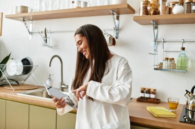 Neşeli genç bir kadın tabletini sıcacık bir mutfakta kullanırken gülümsüyor..