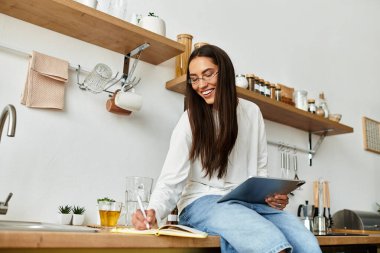 Neşeli genç bir kadın mutfak tezgahına rahatça oturur, elinde kalemle notlar yazar..
