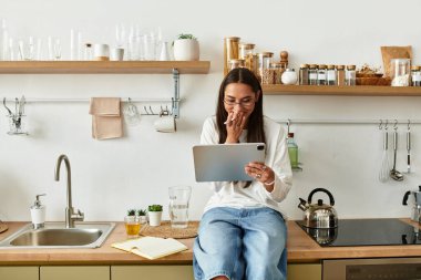 Güzel, genç bir kadın evde vakit geçiriyor, büyüleyici mutfağında oturmuş tableti karıştırıyor..