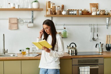 Genç bir kadın düşünceli bir şekilde parlak bir mutfakta durmuş, odaklanmış bir ifadeyle kitap okuyor..