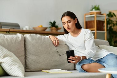 Genç ve güzel bir kadın rahat bir şekilde kanepede oturur, akıllı telefonuyla sade bir şekilde meşgul olur..