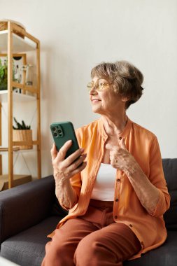 Rahat giysiler içinde, rahat bir oturma odasında akıllı telefonunu kontrol ederken gülümseyen son sınıf öğrencisi bir kadın..