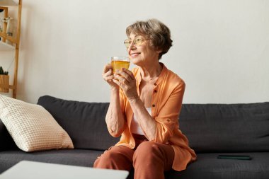 Son sınıf öğrencisi bir kadın kanepede oturuyor, elinde sıcak bir içki, gülümsüyor ve evinde sıcak bir anın tadını çıkarıyor..