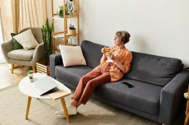 Kıdemli kadın rahat bir şekilde kanepede oturur, bir içki içer, sakin bir öğleden sonranın tadını çıkarır..