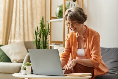 Rahat giyimli son sınıf bir kadın evinde mutlu bir şekilde oturuyor, bilgisayarını neşe ve rahatlıkla kullanıyor..