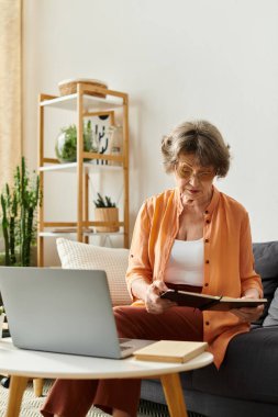 Olgun bir kadın rahat yaşam alanında rahatlar, yakınlarda dizüstü bilgisayarı olan bir kitaba dalmıştır..