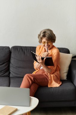 Olgun bir kadın rahat bir şekilde kanepede oturur, huzurlu bir evde gülümseyerek yazar..