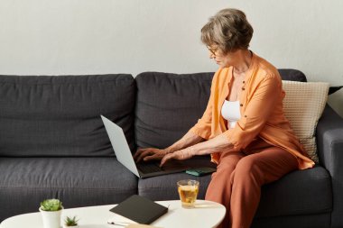 Rahat giyinen son sınıf öğrencisi bir kadın kanepede oturup laptopunu kullanıyor ve sıcak bir içkinin tadını çıkarıyor..