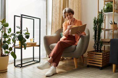 Rahat giyinmiş olgun bir kadın rahat bir sandalyeye oturur ve yakınlarda sıcak bir içecekle yazı yazar..