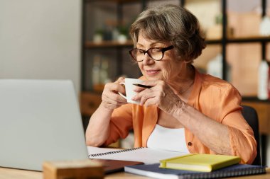 Yumuşak giyimli olgun bir kadın çayını yudumluyor, masasında rahatça oturuyor, çevrimiçi aktivitelerle meşgul..