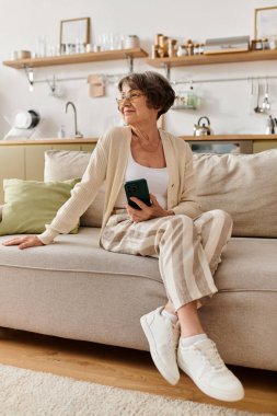 Son sınıf öğrencisi bir kadın, elinde telefonuyla kanepede dinleniyor ve evdeyken rahat bir şekilde giyiniyor..