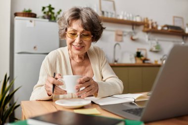 Olgun kadın rahat giysiler içinde rahatlar, çayını yudumlar ve dizüstü bilgisayarına odaklanır..