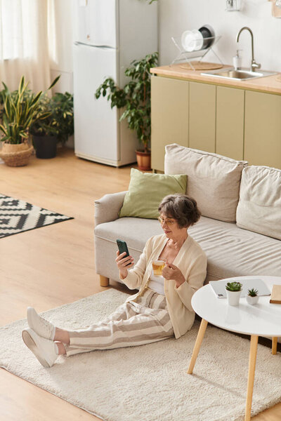 Senior woman relaxes on the floor with a drink, checking her smartphone in a cozy space.