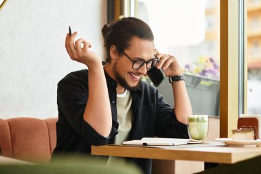 Sakallı adam telefonda gülümsüyor, güneşli bir kafede Matcha ve kekin tadını çıkarıyor..