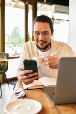 Sakallı genç bir adam kahvesini yudumluyor ve bir kafede dizüstü bilgisayarı üzerinde çalışırken telefonunu kontrol ediyor..