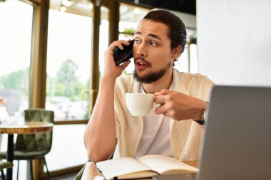 Genç bir adam yaz boyunca hayat dolu bir kafe ortamında kahvesini yudumlarken iş telefonuna cevap veriyor..