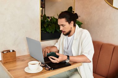Sakallı serbest çalışan biri, hayat dolu bir kafede dinlenirken dizüstü bilgisayarına odaklanıyor..