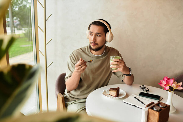 Bearded young man drinking matcha and eating cake in a cozy cafe setting.