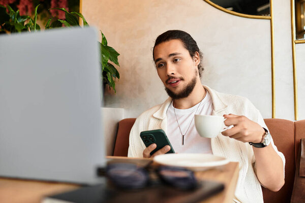 Young man with a beard engages in remote work, balancing tasks on his phone and laptop.