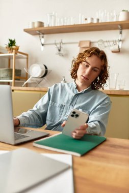 Yakışıklı genç adam ahşap bir masada oturuyor, dizüstü bilgisayarı üzerinde çalışırken akıllı telefonuna dalmış..