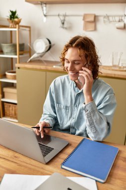 Genç bir adam evde laptopuyla çalışırken telefonda konuşurken oturup gülümsüyor..