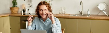 Genç adam telefonda konuşurken gülümsüyor ve evdeki rahat mutfak ortamında notlar alıyor..