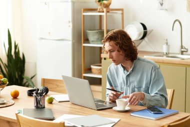 Odaklanmış genç adam rahat bir evde dizüstü bilgisayarıyla çalışırken kahvenin tadını çıkarıyor..